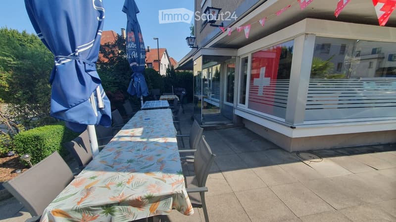 Outdoor patio area with a long table covered in a floral tablecloth, surrounded by chairs and umbrellas, adjacent to a modern building with large windows and a decorative string of flags.
