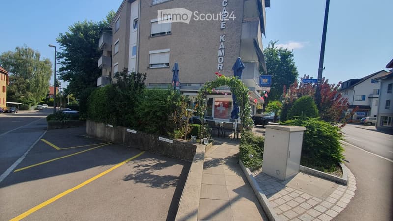 A multi-story building with a sign reading "CAFE KAMOR" on the facade, surrounded by greenery and outdoor seating area.