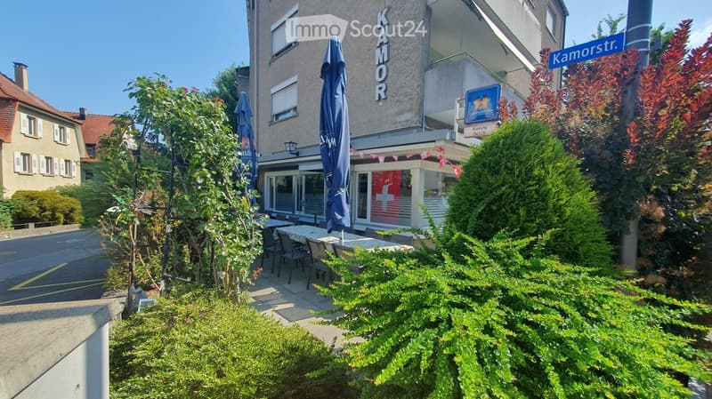 Outdoor seating area with tables and chairs, surrounded by greenery and a building with a sign reading "KAMOR".