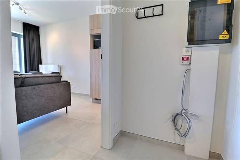 Modern living room with a grey sofa, a small cabinet, and a TV mounted on the wall. The space is well-lit with large windows and neutral-colored walls.