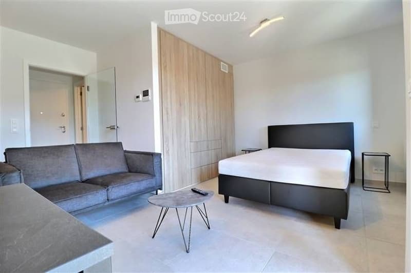 Modern, well-lit room with a grey sofa, a bed with white bedding, a round coffee table, and two nightstands. The room features light-colored walls, a large wooden wardrobe, and a glass door leading to another space.