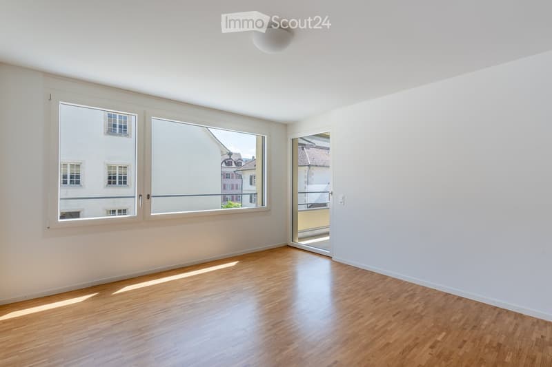 Empty room with wooden flooring, white walls, large windows, and a sliding glass door leading to a balcony.