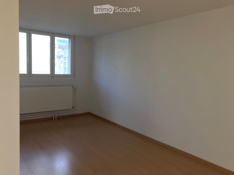 Empty room with wooden flooring, white walls, and large windows allowing natural light. A radiator is visible under the windows.