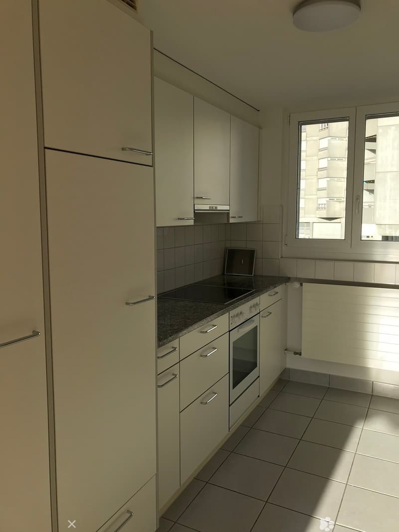 Modern kitchen with light-colored cabinets, a stove, and an oven, featuring tiled floors and a window overlooking a cityscape.