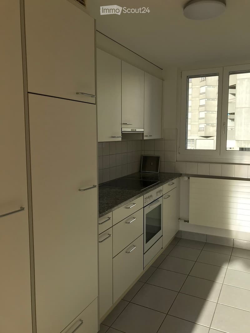 Modern kitchen with light-colored cabinets, a stove, and an oven, featuring tiled floors and a window overlooking a cityscape.