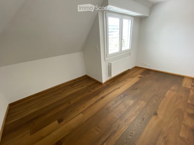 Empty attic room with wooden flooring, white walls, and a single window with a radiator below it.