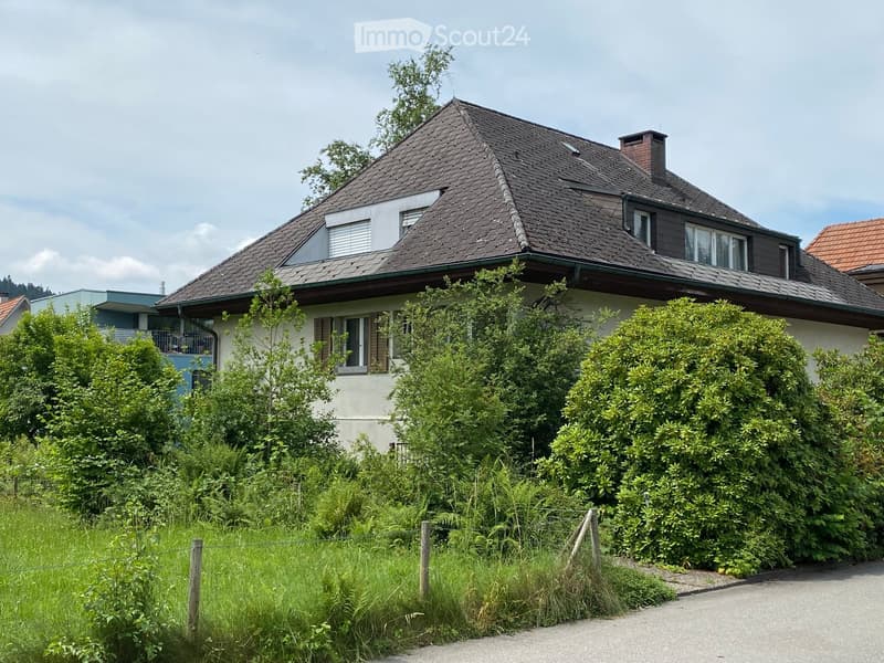 Two-story house with a dark tiled roof, multiple windows, and a chimney. The house is surrounded by greenery and has a fenced front yard.