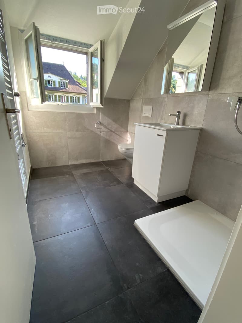 Modern bathroom with dark tiled floor, white sink, toilet, bathtub, and large mirror. Large window offers view of the outside.