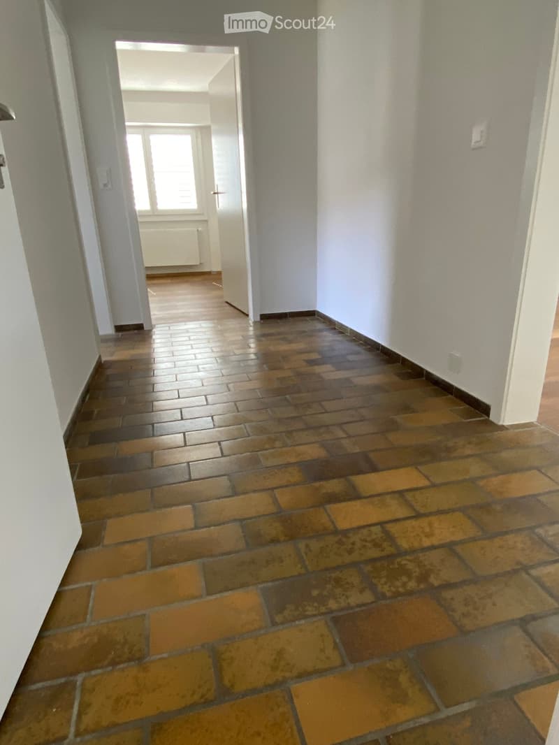 Empty hallway with tiled floor and white walls, leading to an open room with a window and radiator.