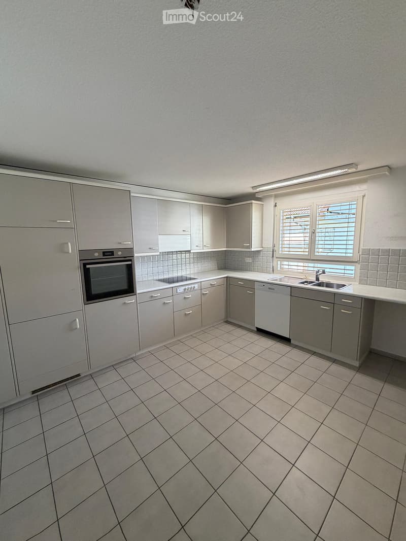 Modern kitchen with grey cabinets, tiled floor, built-in oven, sink, and dishwasher. Natural light from a window with blinds.
