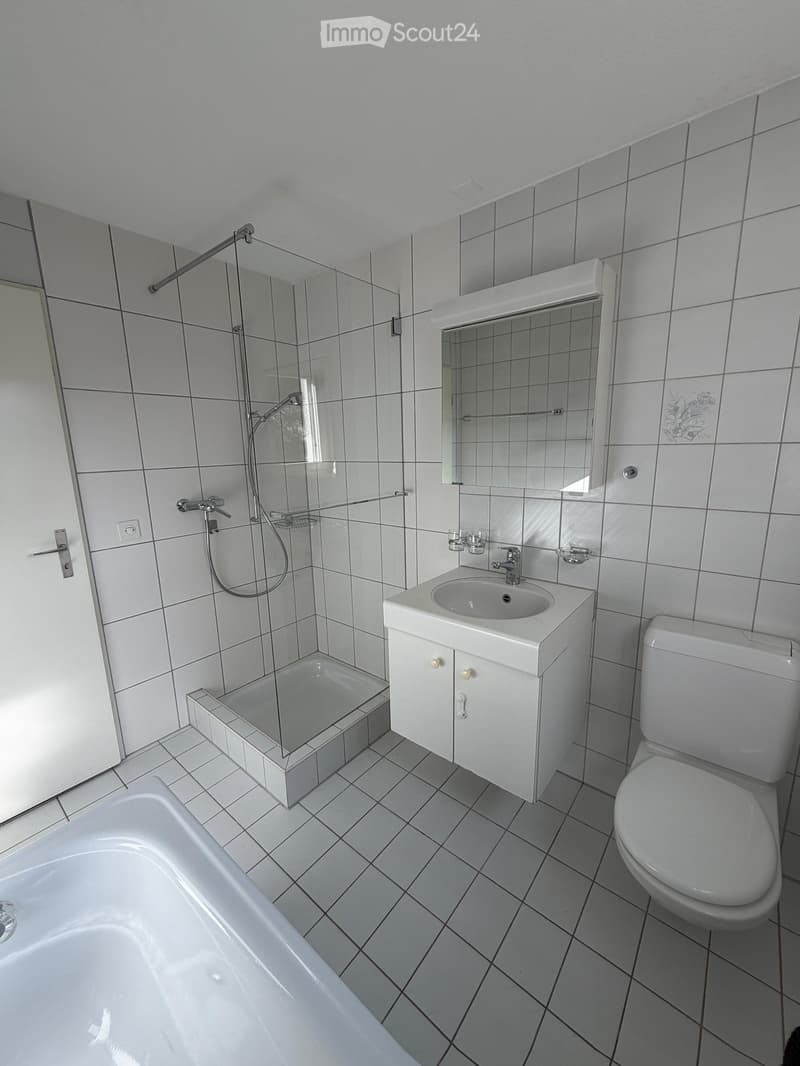 Modern bathroom with white tiles, bathtub, toilet, sink, mirror, and glass-enclosed shower area.