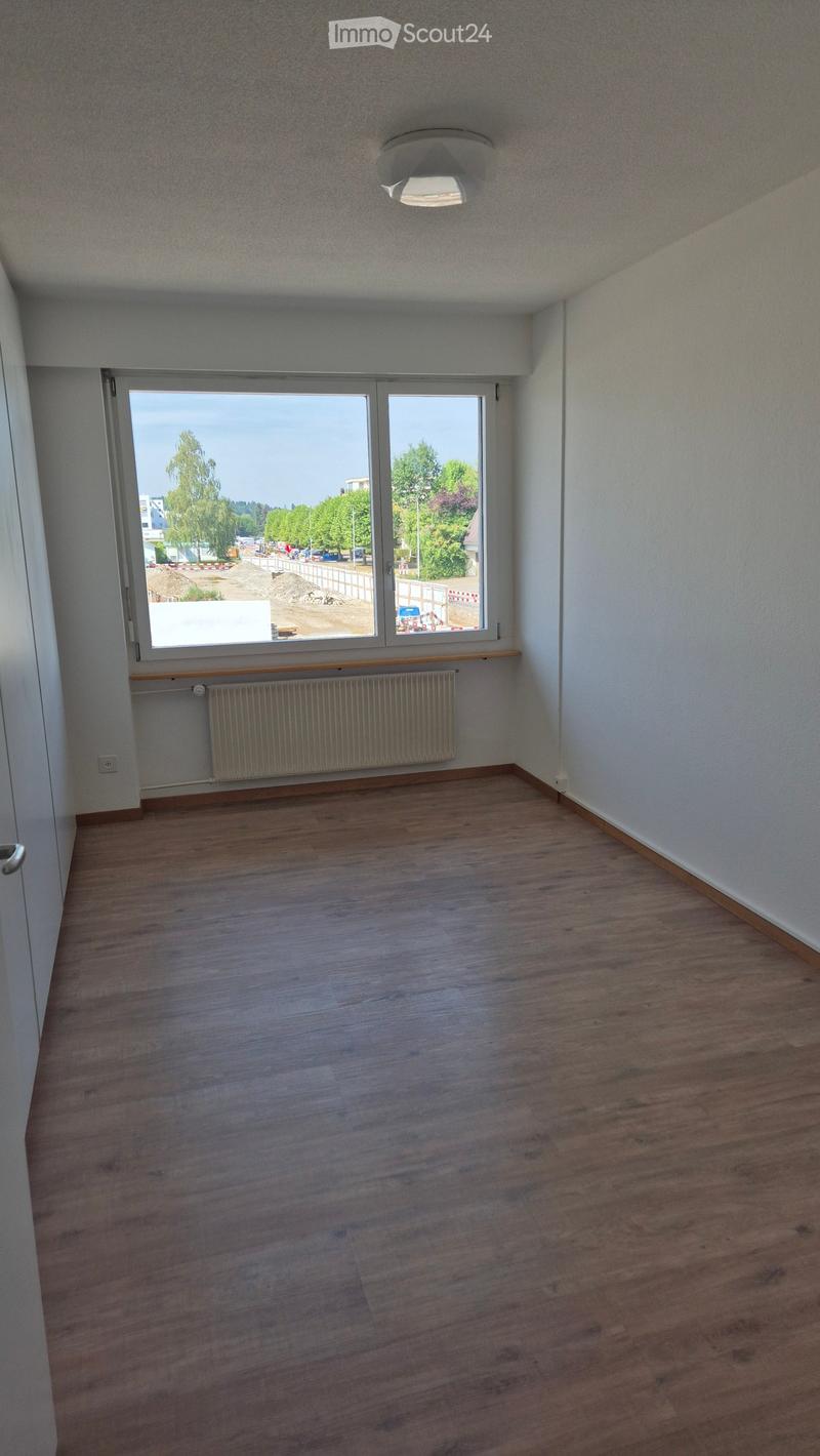 Empty room with wooden flooring, white walls, a radiator, and a large window showing a view of trees and a construction site.