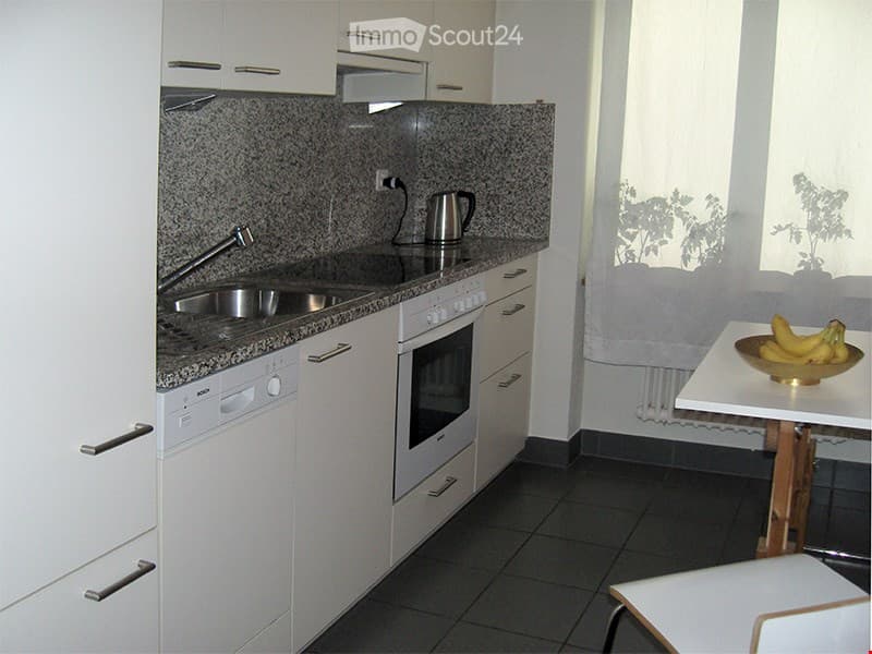 Modern kitchen with white cabinets, granite countertop, stainless steel sink, built-in oven, dishwasher, and a small dining area with a table and chairs.