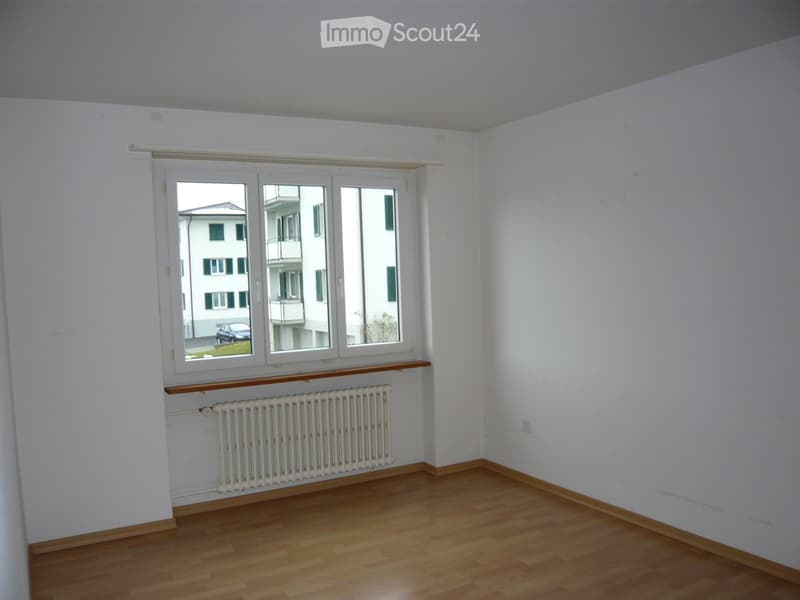 Empty room with light wooden flooring, white walls, a large window with white frame, and a white radiator below the window.