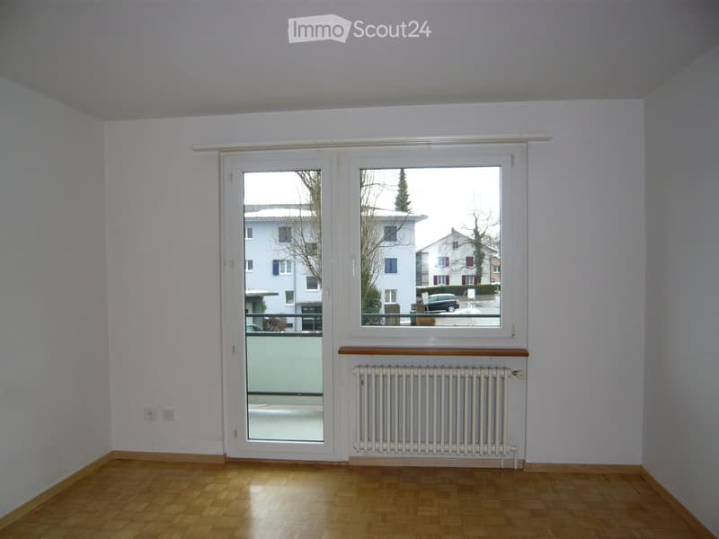 Empty room with wooden flooring, white walls, large window, radiator, and sliding glass door leading to a balcony.