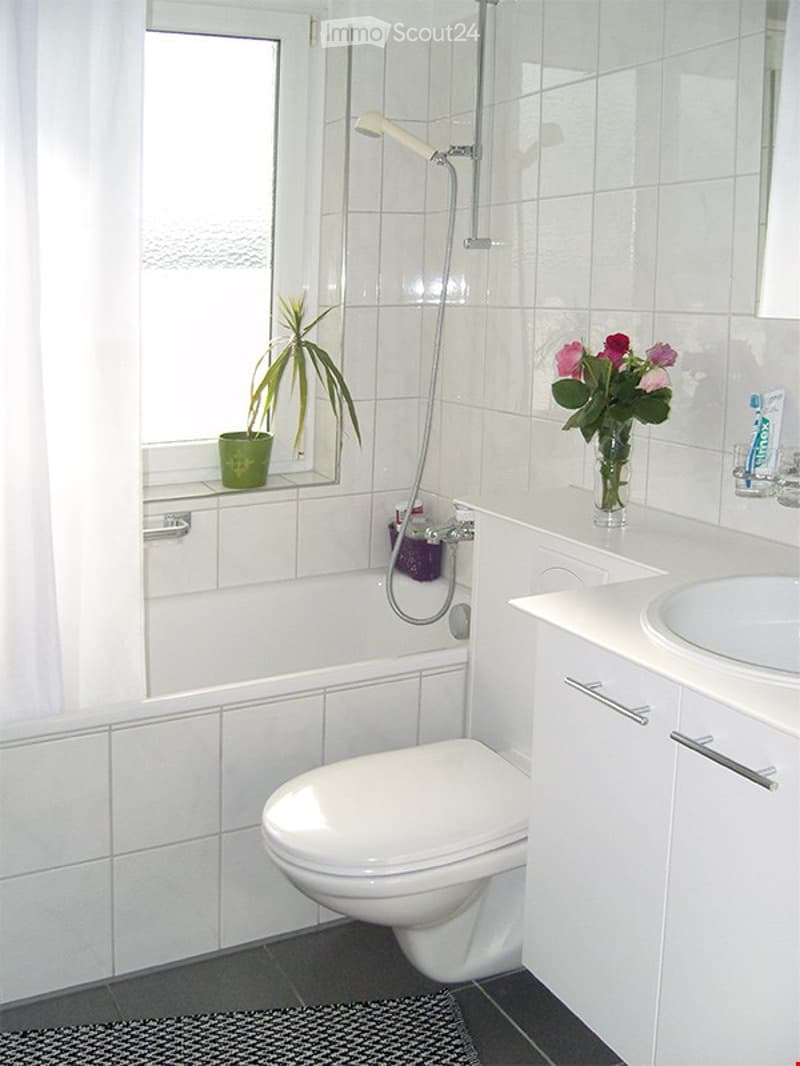 Modern bathroom with white tiles, a toilet, sink, bathtub with a shower, a small plant on the window sill, and a vase with flowers on the counter.