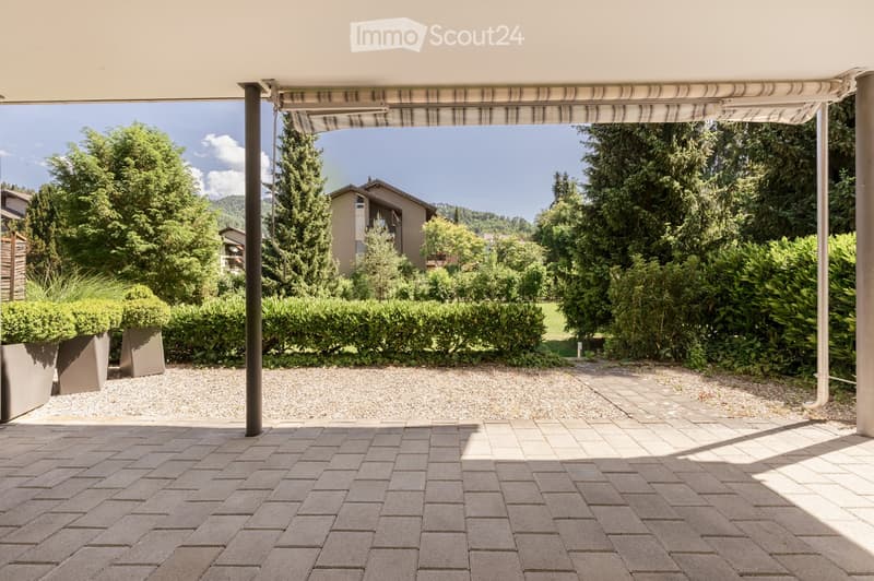 A covered outdoor patio with brick tiles, gravel area, greenery, and a view of a house and trees in the background.