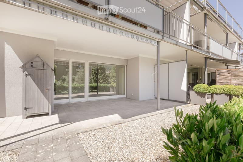 Courtyard area with gravel ground, small wooden shed, potted plants, and a modern building with large glass windows and a balcony.