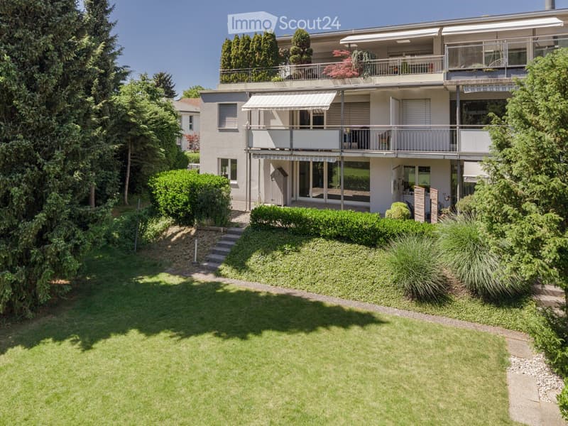 Modern two-story house with balconies, surrounded by a well-maintained garden with green lawns, shrubs, and trees.