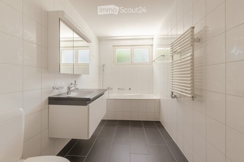 Modern bathroom with white tiles, black countertop, sink, bathtub, mirror, radiator, and two small cups on the countertop.