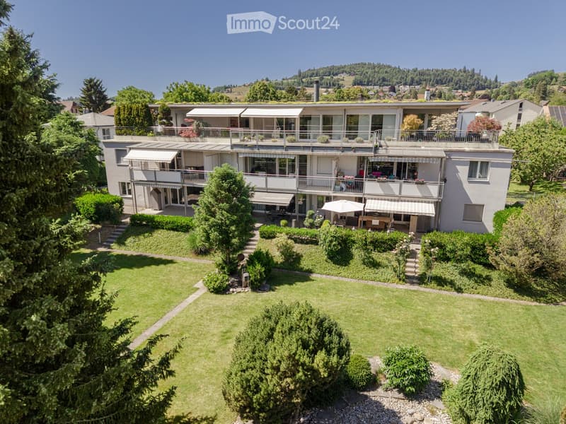 A modern two-story house with large windows, balconies, and a well-maintained garden with a lawn, shrubs, and trees.