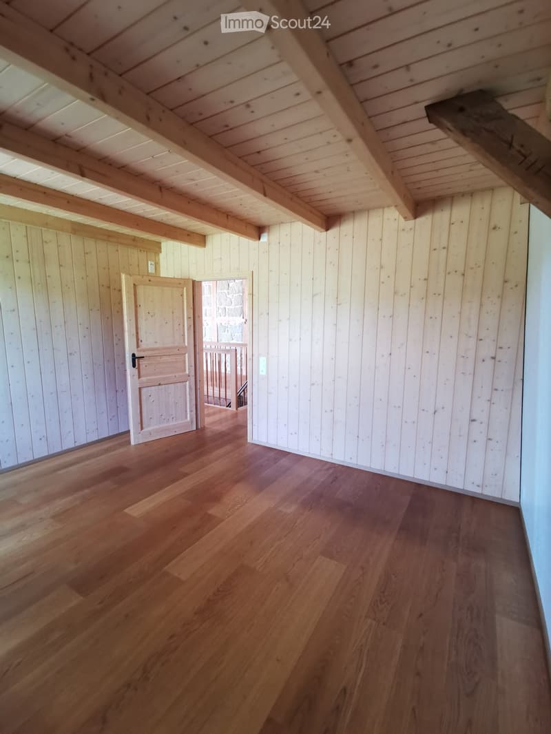 Empty room with wooden walls, wooden floor, and exposed wooden beams on the ceiling. A door leads to another area with a stone wall visible.