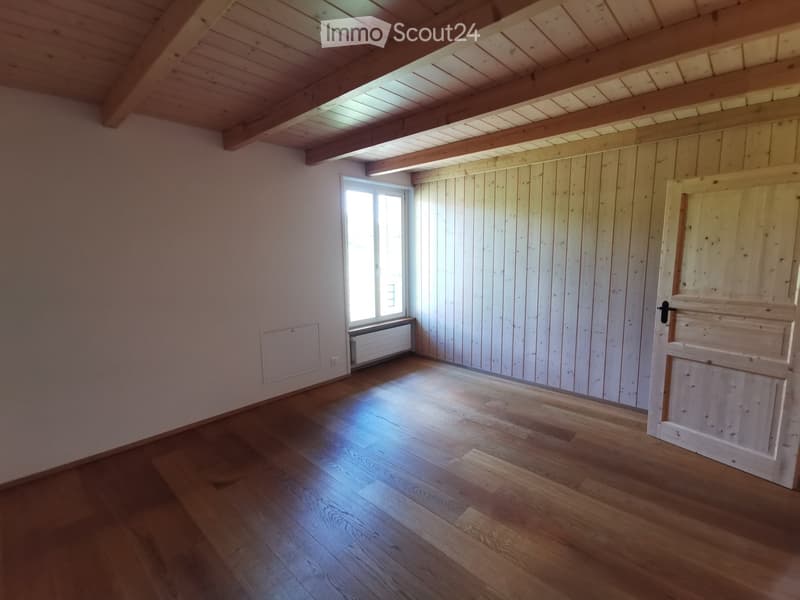 Empty room with wooden flooring, exposed wooden beams on the ceiling, white paneled walls, and a single window letting in natural light.