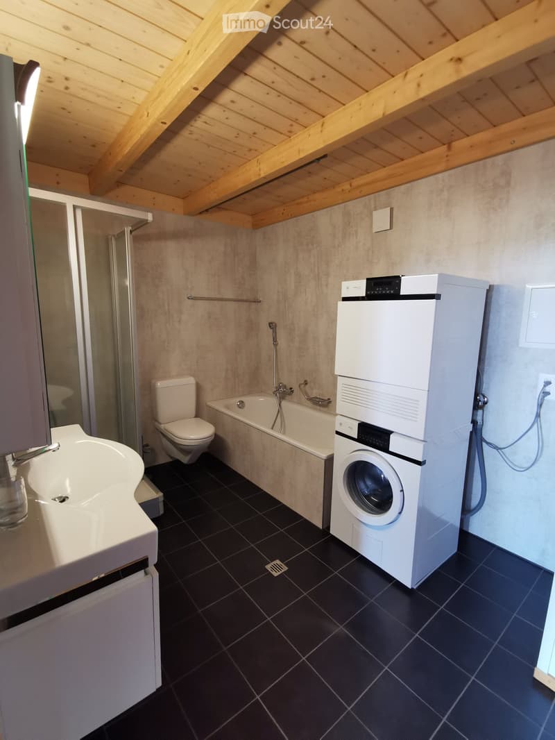 Modern bathroom with dark tiled floor, white sink, toilet, bathtub, and stacked washing machine/dryer unit. Features exposed wooden beams on the ceiling and a glass-enclosed shower area.