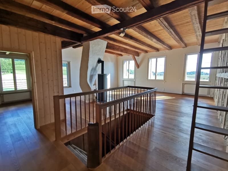 Open, airy space with wooden floors and exposed wooden beams on the ceiling. Large windows provide natural light, and a staircase leads to an upper level.