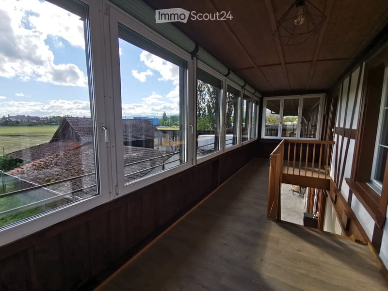 Spacious balcony with wooden flooring, large windows offering a view of the countryside, and a wooden railing leading to an outdoor area.