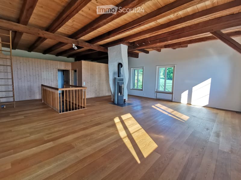Empty room with wooden floors, exposed wooden beams, white walls, and a staircase leading to another level. There is a wood-burning stove in the center.