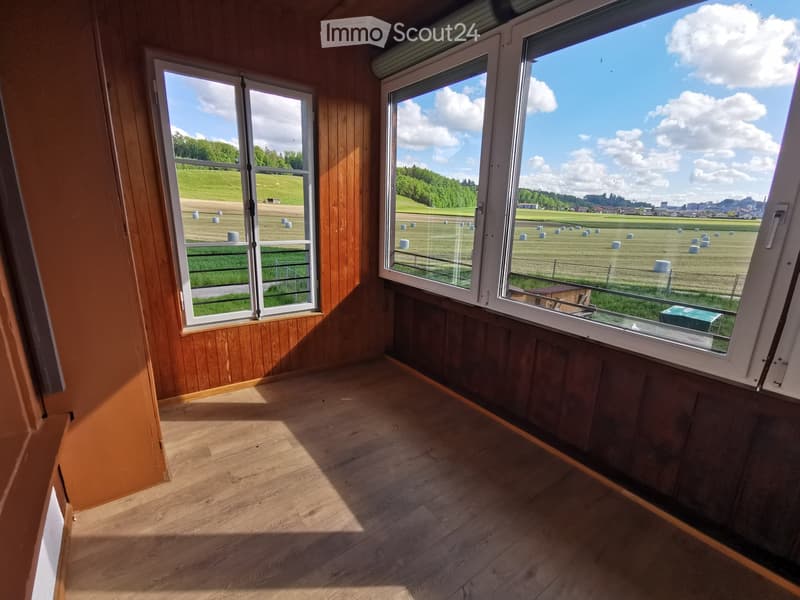 Empty room with wooden walls and floor, large windows offering a view of a green field with hay bales.