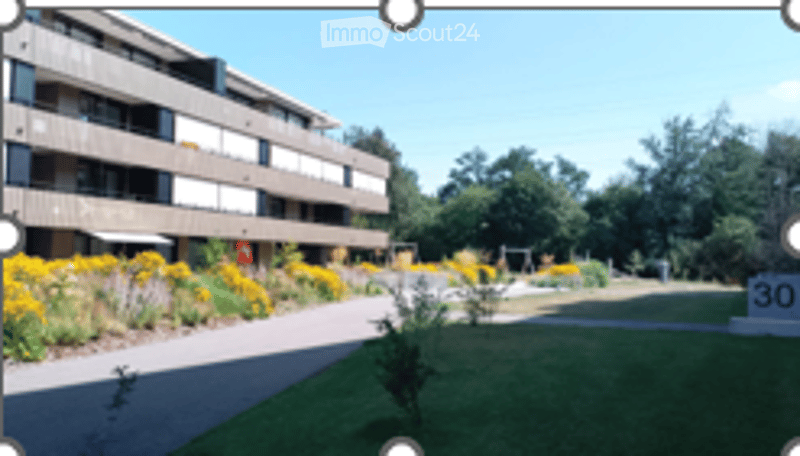 Modern multi-story apartment building with balconies and surrounding greenery, including a well-maintained lawn and flower beds.