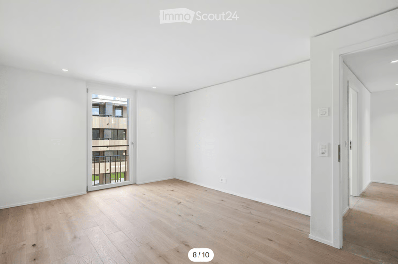 Empty room with white walls, wooden flooring, and a large window with a view of a building outside.