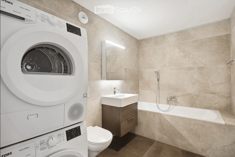 Modern bathroom with beige tiled walls, a white washing machine, a sink with a mirror, a toilet, and a bathtub with a handheld shower.
