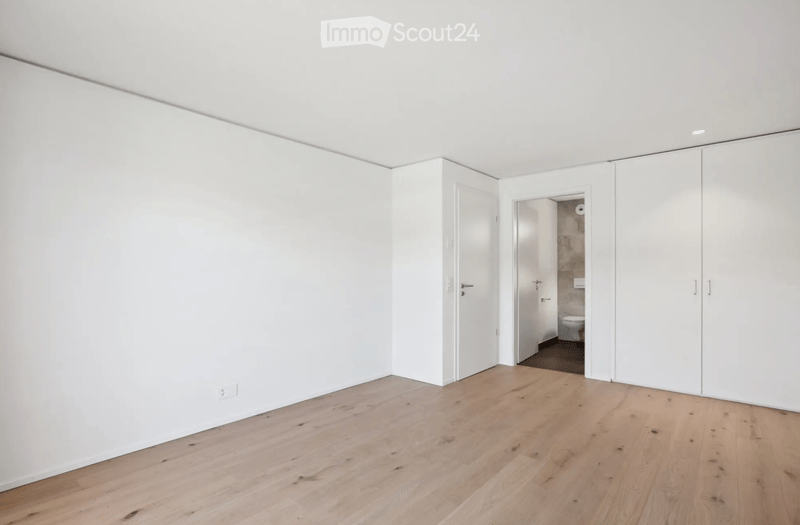 Empty room with white walls, light wooden flooring, and built-in white closet units. A doorway leads to a small bathroom with a toilet and stone wall.