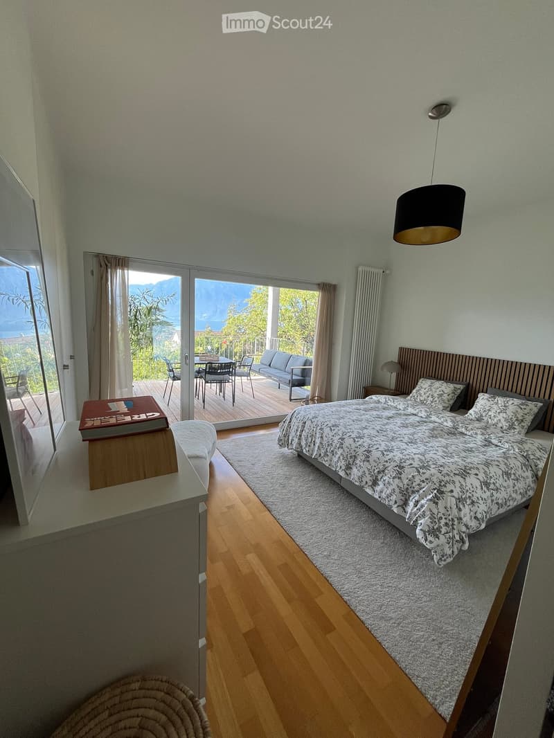 Bright bedroom with wooden flooring, large bed with patterned bedding, sofa, dining table and chairs on a balcony, and a view of the mountains.