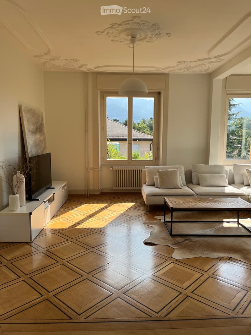 Elegant living room with a geometric parquet floor, large windows offering a scenic mountain view, a modern sofa set, a sleek coffee table, and a TV stand with a screen.