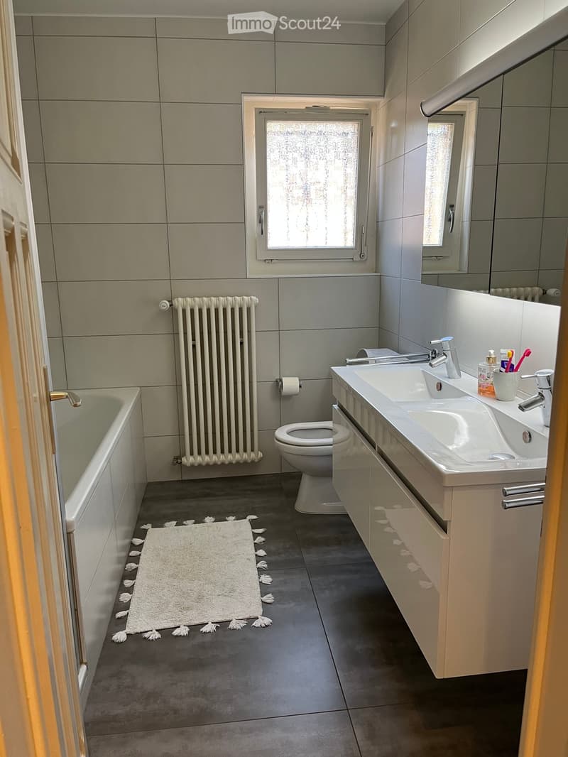 Modern bathroom with a bathtub, toilet, double sink, and a large mirror. Features a white radiator, tiled walls, and a white bath mat on the floor.