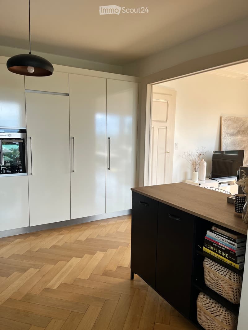 Modern kitchen with white cabinets, wooden floor, island counter, and a view to a dining area through an open doorway.