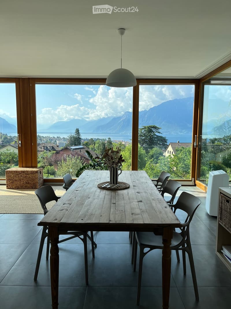Bright kitchen with large windows offering a scenic mountain view, featuring a wooden dining table, chairs, and a decorative vase.