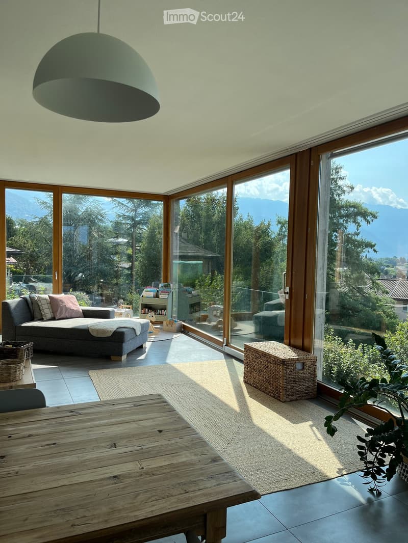 Bright living room with large glass doors offering a scenic mountain view, featuring a grey sofa, wooden table, wicker basket, bookshelf, and a pendant light.