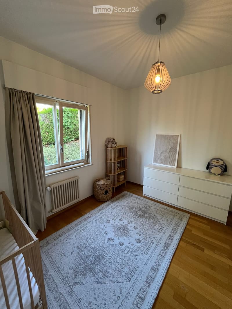 Brightly lit room with wooden flooring, large window with curtains, white dresser, wooden shelf, wicker basket, rug, and decorative items.