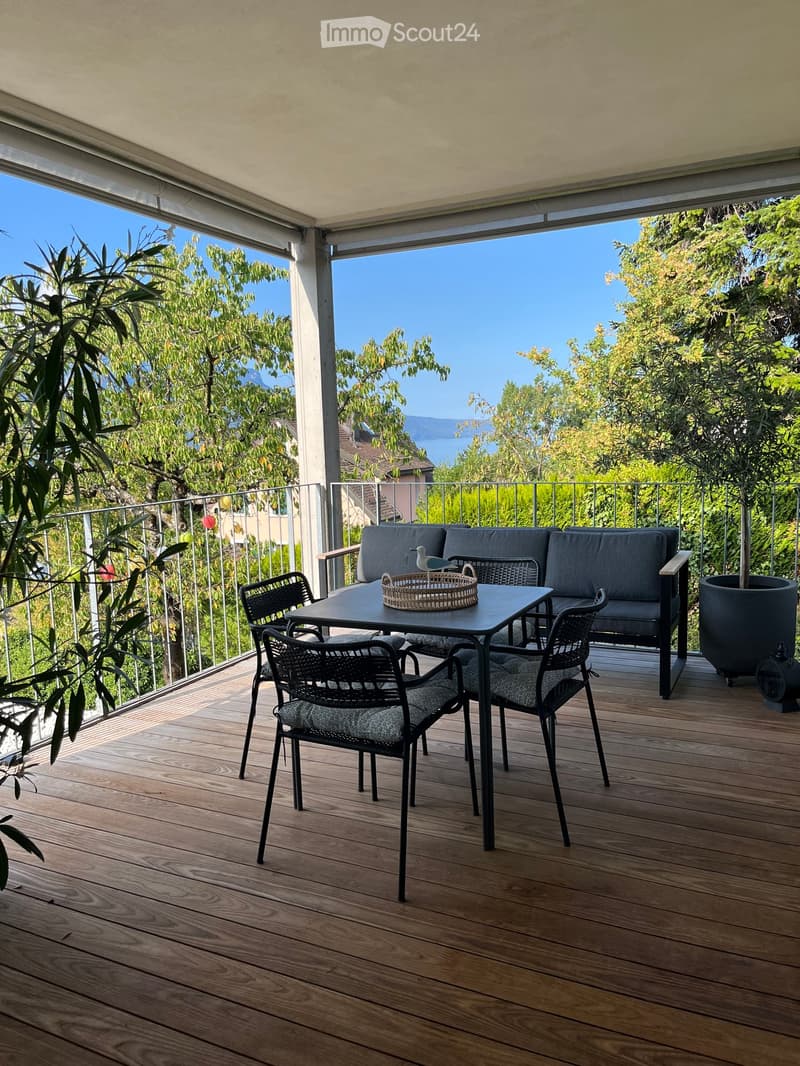 Wooden deck with a sofa, table, chairs, and a wicker basket, overlooking a scenic view of trees and a lake.