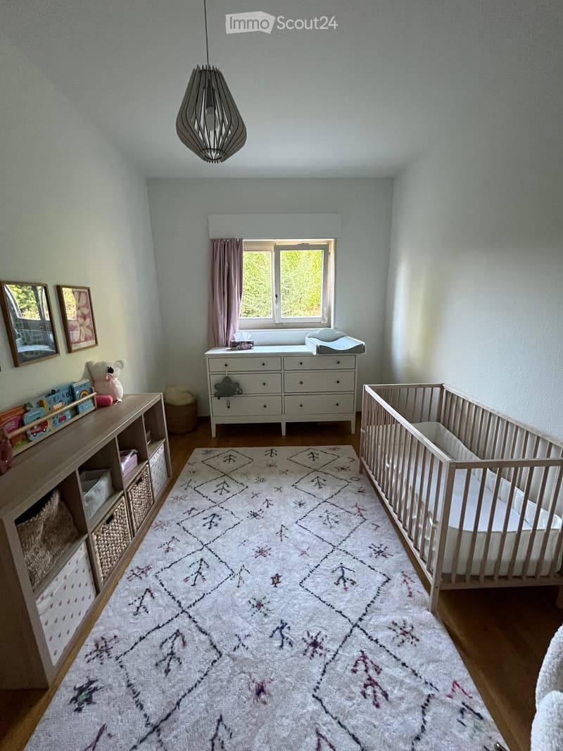 Brightly lit children's bedroom with a crib, dresser, shelf with toys, and a patterned rug.