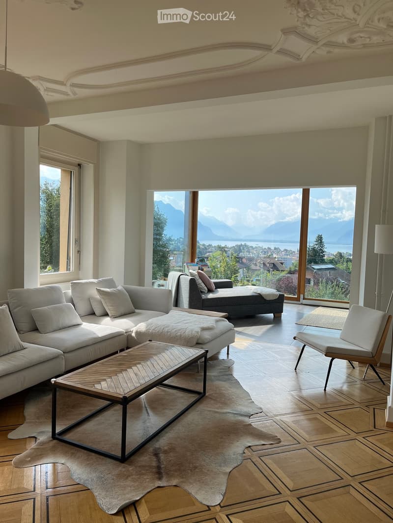 Modern living room with large windows offering a scenic mountain view, featuring a grey sectional sofa, a wooden coffee table, a white armchair, and an ottoman.