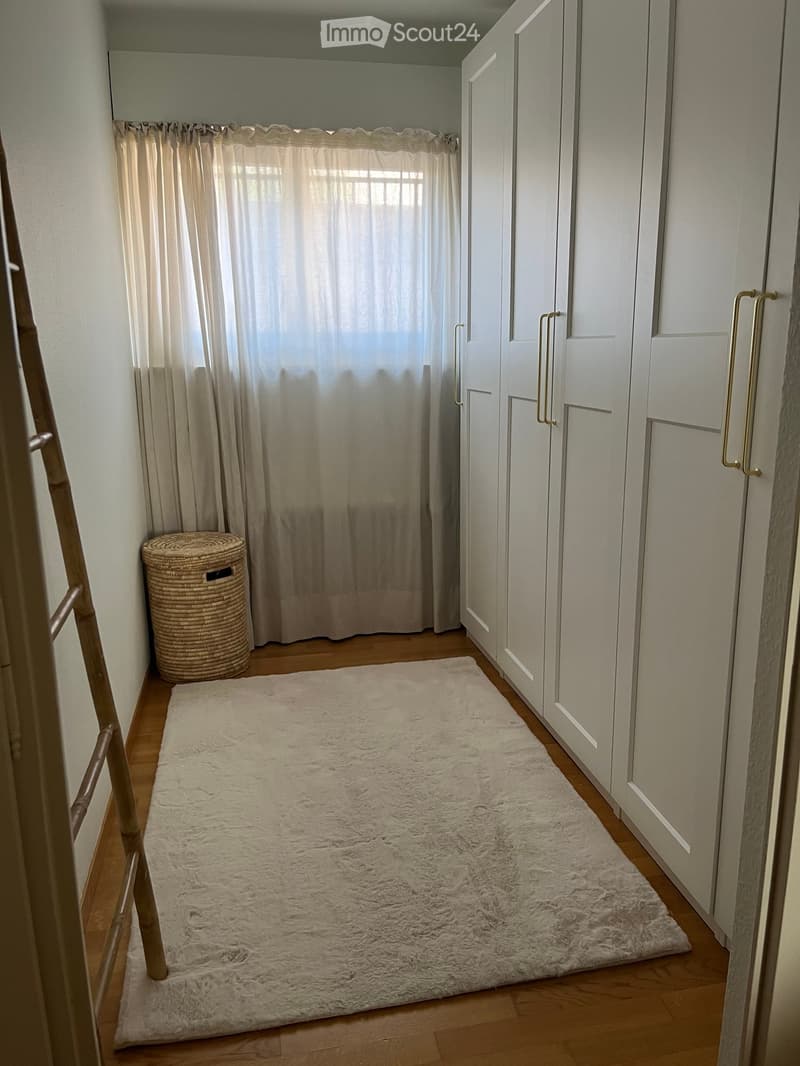 A narrow hallway with light wooden flooring, white walls, and built-in white closet units with silver handles. A large, fluffy white rug lies on the floor, and a woven basket sits near the window with sheer curtains.