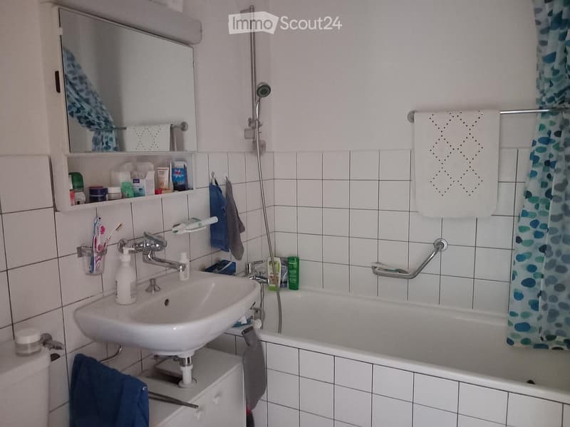 A small bathroom with a white tiled wall, a sink with a faucet, a toothbrush holder with toothbrushes, a mirror above the sink, a bathtub with a shower curtain, and a towel hanging on the side.