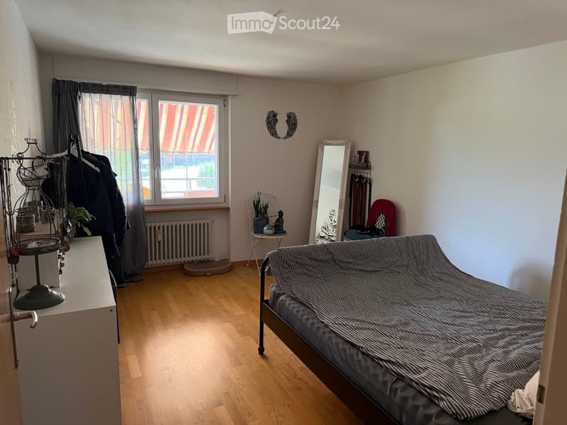 A small, well-lit bedroom with wooden flooring, a grey sofa, a large mirror, a small table with a plant, and a radiator under the window.