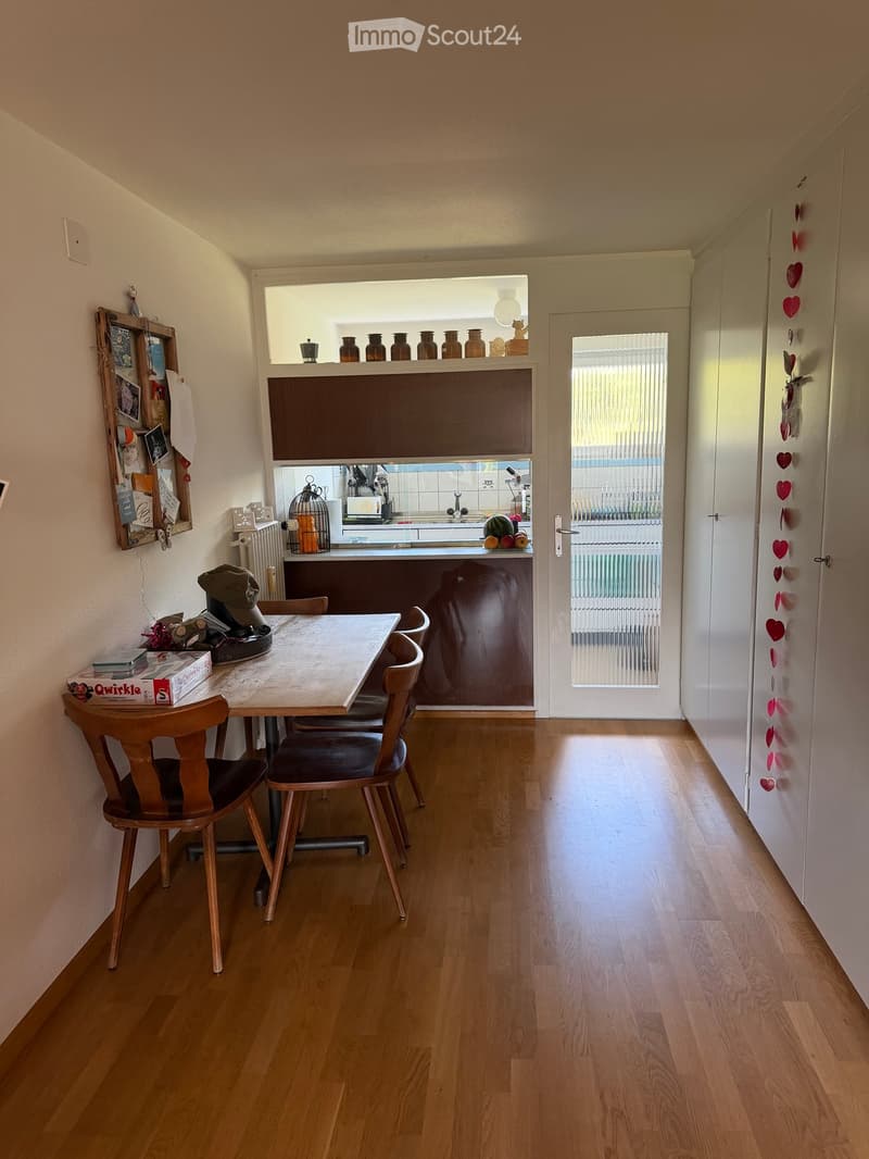 A small, well-lit room with wooden flooring, a dining table with chairs, a small kitchen area visible through an open doorway, and various decorative items including vases and a birdcage.